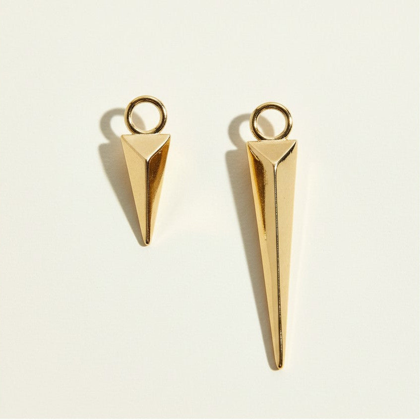 This pair of solid gold charms features a faceted triangular spike silhouette, tapering from a flat pyramid top into a sharp, elongated point for a bold, architectural look. Each charm is topped with a round jump ring for easy attachment to necklaces, bracelets, or hoop earrings, and the set includes two proportional sizes — a shorter charm and a longer, more dramatic drop — allowing for mixed-length layering. 