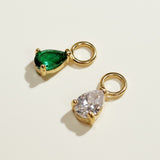 These interchangeable earring charms feature a pear-cut crystal set in a four-prong gold setting, topped with a round jump ring for easy attachment to hoops or huggies. Available in two colorways — a vivid emerald green and a sparkling clear crystal — the charms let shoppers customize and mix up their earring stack with a single pair of base hoops. Photographed flat against a clean, neutral background, the dramatic side lighting highlights each stone's faceted cut and the polished gold prongs, along with th