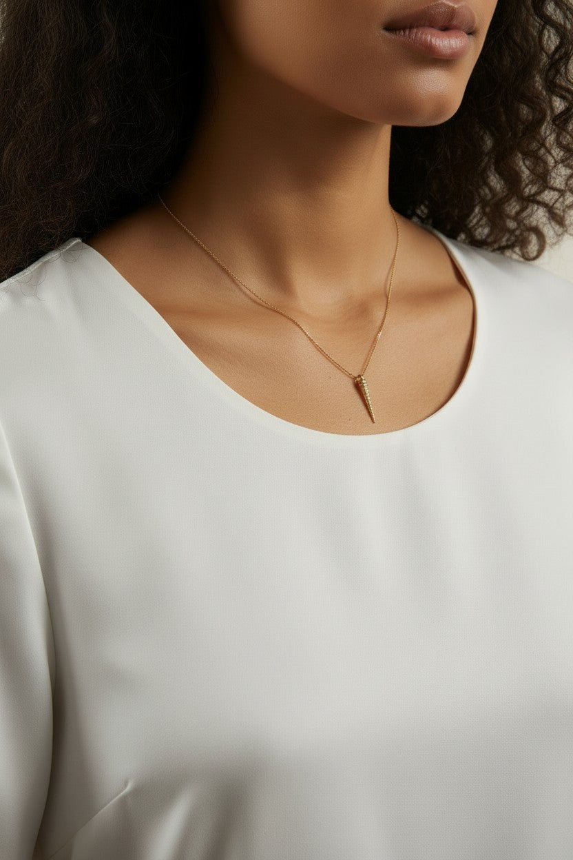 This close-up shot shows a gold dagger pendant resting just below the collarbone on a fine box chain, styled with a round scoop-neck white top for a soft, minimal presentation. The pendant's crystal-lined taper and sharp point are shown at true scale against warm skin, giving shoppers a clear sense of its delicate drop length and dainty proportions.