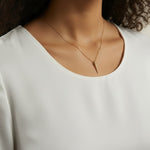 This close-up shot shows a gold dagger pendant resting just below the collarbone on a fine box chain, styled with a round scoop-neck white top for a soft, minimal presentation. The pendant's crystal-lined taper and sharp point are shown at true scale against warm skin, giving shoppers a clear sense of its delicate drop length and dainty proportions.