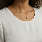 This close-up shot shows a gold dagger pendant resting just below the collarbone on a fine box chain, styled with a round scoop-neck white top for a soft, minimal presentation. The pendant's crystal-lined taper and sharp point are shown at true scale against warm skin, giving shoppers a clear sense of its delicate drop length and dainty proportions.