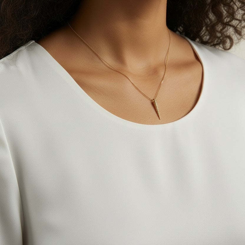 This close-up shot shows a gold dagger pendant resting just below the collarbone on a fine box chain, styled with a round scoop-neck white top for a soft, minimal presentation. The pendant's crystal-lined taper and sharp point are shown at true scale against warm skin, giving shoppers a clear sense of its delicate drop length and dainty proportions.