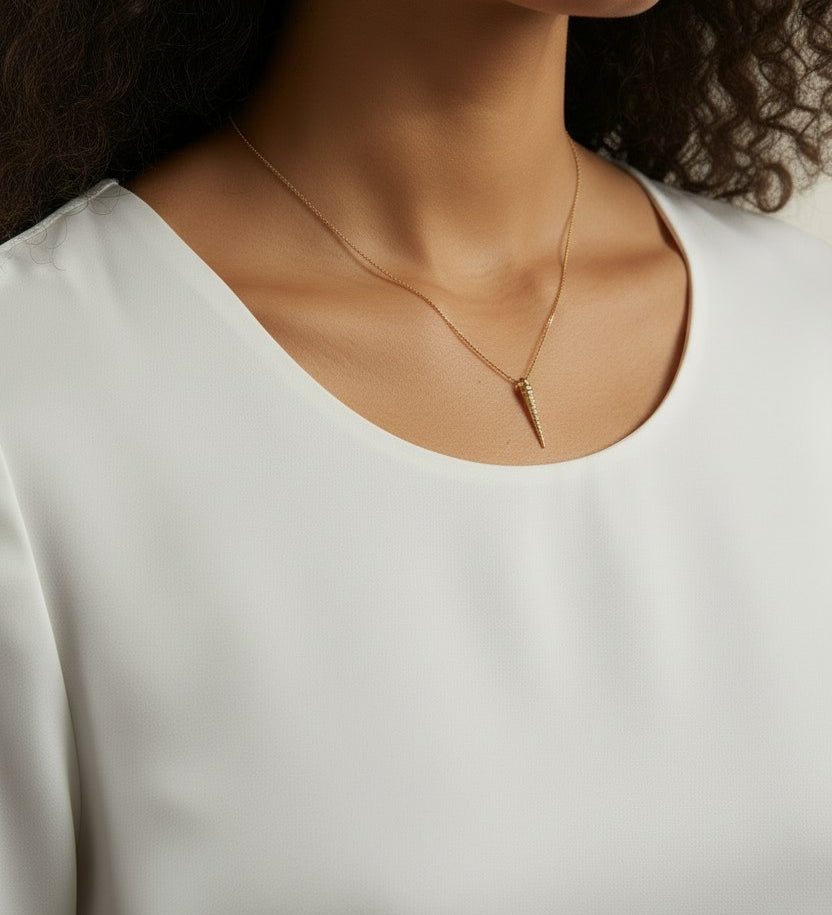 This close-up shot shows a gold dagger pendant resting just below the collarbone on a fine box chain, styled with a round scoop-neck white top for a soft, minimal presentation. The pendant's crystal-lined taper and sharp point are shown at true scale against warm skin, giving shoppers a clear sense of its delicate drop length and dainty proportions.